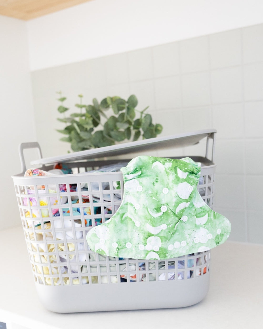 A white plastic laundry basket with a lid, filled with colorful folded items and topped with a green and white patterned cloth, set on a white surface against a white tiled wall.